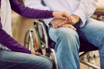 A woman sitting with her hand on top of a man's hands. The man is in a wheelchair.