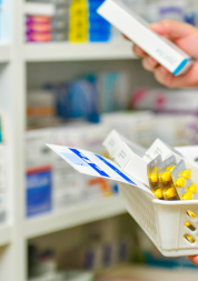 Close up showing a pharmacist's hands and arms. They are filling a prescription basket, standing in front of shelves containing medication boxes.