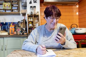 A woman sitting at a table in a kitchen, looking at a mobile phone and writing on a notepad.